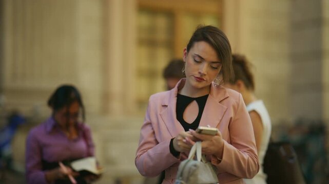 A confident young businesswoman engages in a phone call and uses her smartphone on a bustling city street. She interacts with her mobile device in an urban outdoor setting.