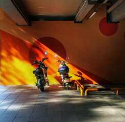 Two parked scooters illuminated by dramatic sunlight in an urban parking area. Bold orange and red geometric wall patterns, diagonal sunbeams, and contrasting shadows create a striking interplay of co © Liudmila Dmitrieva