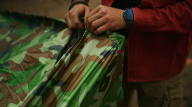 Man pitches a camouflage tent in a pine forest, threading poles through fabric sleeves during outdoor camping preparation.