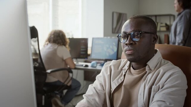 African American man wearing glasses analyzing project on computer monitor in office, rack focus shot. Useful for tech startup advertising, software engineering training, and corporate recruitment