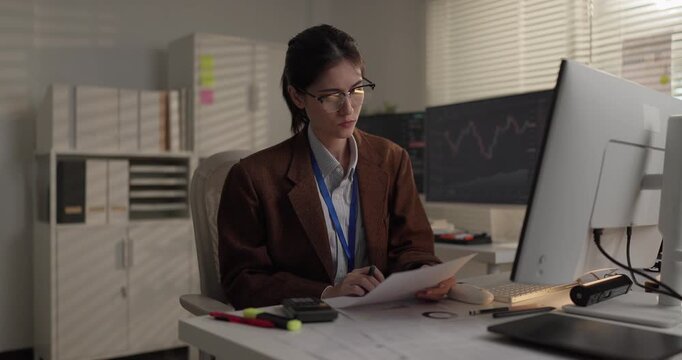 Woman stock trader writing notes and reviewing printed report at trading desk in office