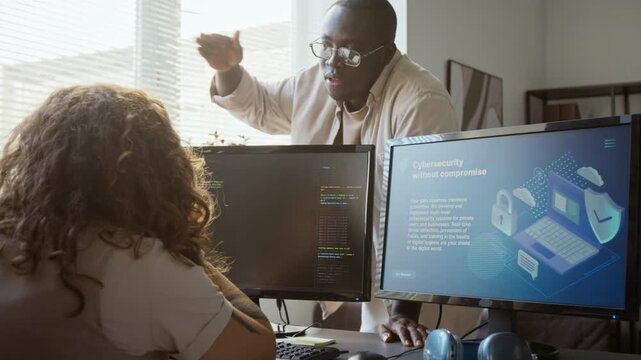 African American young adult software engineer gesturing while explaining cybersecurity code to female colleague at computer monitors. Clip suits IT training and cybersecurity campaigns