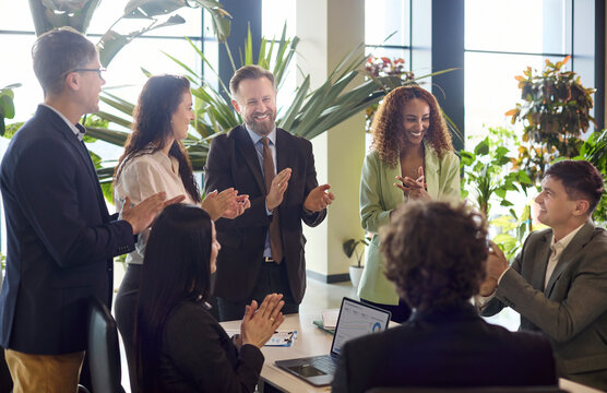 Business team meeting in office with applause success. Coworkers clap around a table in a bright workspace with plants and laptops. Clear teamwork achievement concept for business branding.