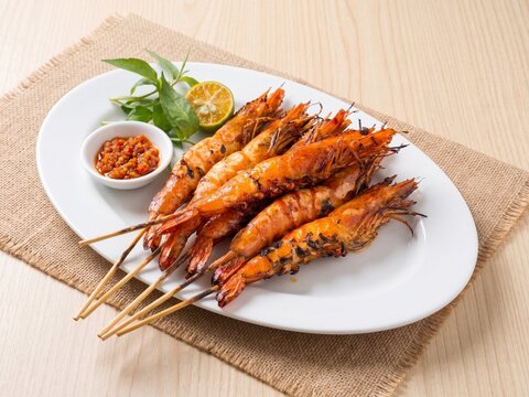 Grilled skewered jumbo prawns accompanied by sambal chili paste, lime, and herbs arranged on a rustic sackcloth runner.