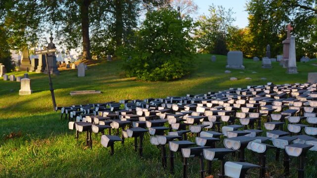 Rows of small lights fill a cemetery lawn beside grave markers. White lenses face across green grass beneath trees with a cross monument on the right.