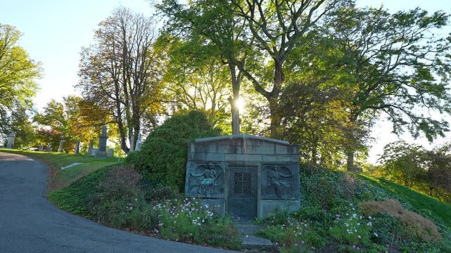 A mausoleum sits in a planted slope beside a curving road. Sunlight filters through trees above relief panels and flowers surrounding a gray entrance.