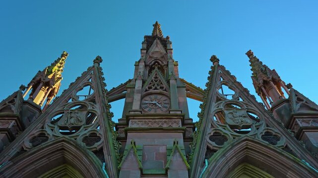 Pointed gables frame the central pinnacle of a Gothic cemetery gate. Brown masonry and green surface stains border open tracery against a blue sky.
