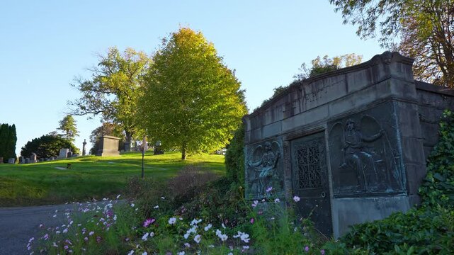 A stone tomb sits beside a path and flowering plants. Winged reliefs flank a metal door while trees rise above the gray facade in sunlight.