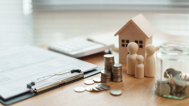 Wooden house and family figures beside stacked coins, a savings jar and financial documents on a desk, wealth planning, emergency savings and long-term family security.