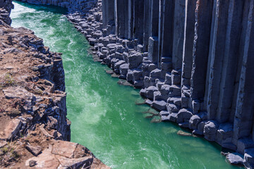 A green river flows below towering basalt columns in Studlagil canyon, East Iceland. Sunlight catches the moving water beside dark cliffs and a rough, rocky foreground. © Artem