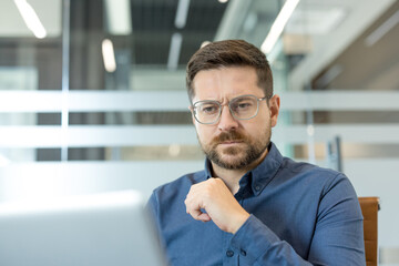 Naklejka na ściany i meble Focused professional sits at desk in bright office, looking at laptop with thoughtful expression. He appears to review information, consider decisions, and plan work tasks carefully.