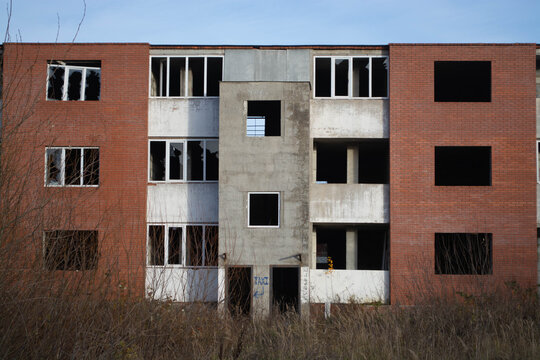 Unfinished brick and concrete building with empty window openings amid overgrown weeds. Uncompleted construction site. Urban decay and stalled development. Building industry