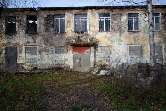 Dilapidated two-story building with peeling plaster and broken windows beside an overgrown yard. Urban decay and structural neglect. Abandoned architecture, disrepair and deterioration