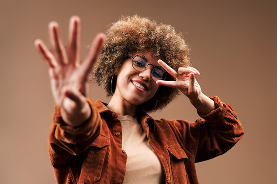 Smiling young woman with curly afro hair and glasses posing against a warm brown studio background, raising her hands in a playful gesture while wearing a rust corduroy jacket.