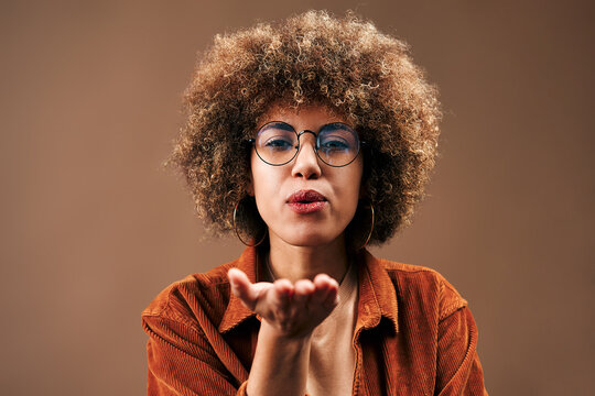 Close portrait of a confident woman with curly hair and glasses blowing a kiss toward the camera against a warm brown background.