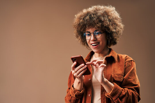 Smiling young woman with curly hair and glasses using a smartphone against a warm brown studio background with copy space.
