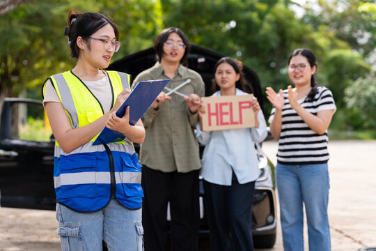 A female roadside assistant records details on a clipboard in front of a car with its hood open while three women stand nearby, conveying vehicle trouble, support service, and teamwork.
