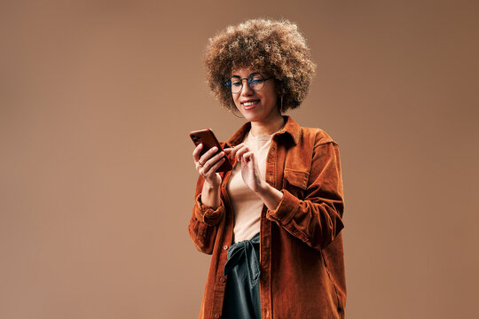 Smiling young woman with curly hair and glasses uses a smartphone in a warm studio portrait, wearing a brown jacket against a simple beige background.