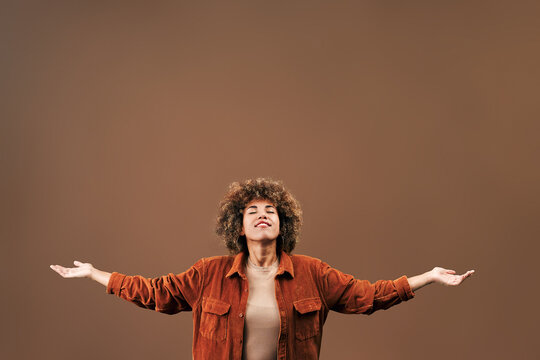 Joyful young woman with curly hair standing against a warm brown background, smiling with arms open in a welcoming studio portrait with copy space overhead.