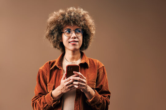 Joyful young woman with curly afro hair and glasses looking up with a curious smile while holding a smartphone against a warm beige studio background.