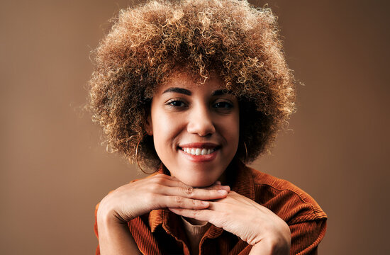 Smiling young woman with curly hair resting her chin on her hands against a warm brown studio background