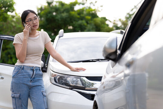 Asian woman calls on her phone while resting a hand on a white car beside another vehicle, portraying possible accident reporting, auto insurance claims, and roadside assistance.