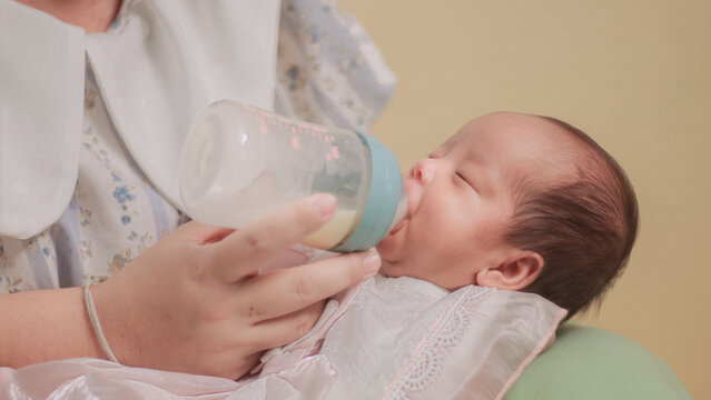 Infant feeding, daily childcare, nutrition, and nurturing. Newborn baby drinking milk from a feeding bottle held by an adult, mother held him in her lap. Soft close up with pastel bedding.