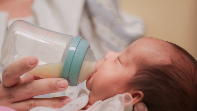 Infant feeding, daily childcare, nutrition, and nurturing. Newborn baby drinking milk from a feeding bottle held by an adult, mother held him in her lap. Soft close up with pastel bedding.