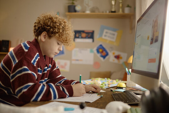 Caucasian teenage boy studying online with remote tutor on computer screen, writing notes during distance learning class. Concept for edtech platforms and online tutoring service advertising