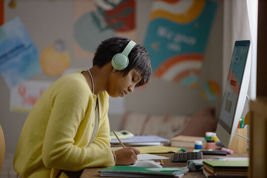 African American teenager with headphones writing notes near computer screen during online tutoring lesson. Perfect for elearning campaigns and remote high school education concepts