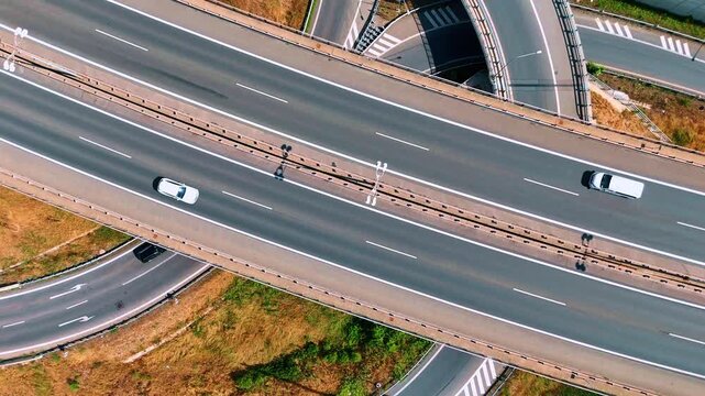 A divided highway crosses above curved access ramps. White vehicles and dashed lane markings contrast with the gray asphalt viewed from above.