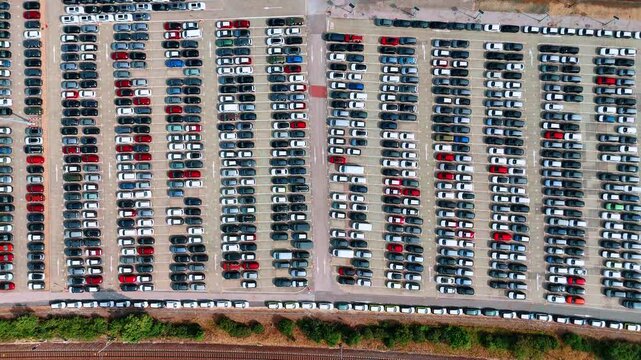 Parked cars form parallel rows across a storage yard. A vertical view shows white and dark vehicles beside a railway along the lower edge.