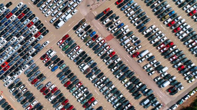 Diagonal rows of cars cover a paved vehicle storage lot. White and dark roofs repeat between empty aisles in a steep aerial view.