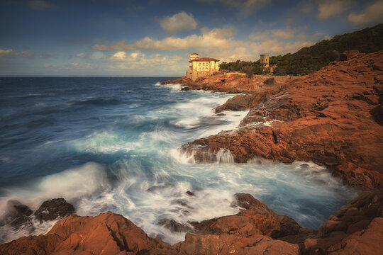 Livorno, Italy - 02 October 2019: View of Scogli Piatti C 7 with its historic villa on the rugged red rocky coastline, surrounded by crashing turquoise waves under a dramatic sky.