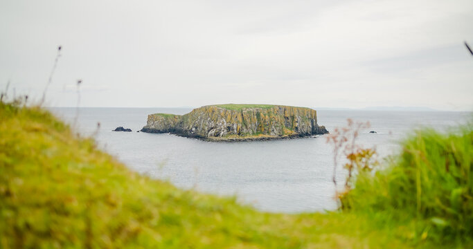 Ballintoy, County Antrim, United Kingdom - 14 September 2024: View of Carrick-a-Rede island surrounded by the Atlantic Ocean with grassy cliffs in the foreground.