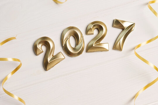 Close up of large golden 2027 numerals tilted diagonally with gold ribbons on white wooden surface