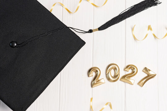 Black academic cap with stretched tassel next to gold 2027 numbers and ribbon on white wooden table