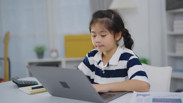 Young girl using laptop computer indoors at home with notebook calculator and chart paper nearby focused on screen