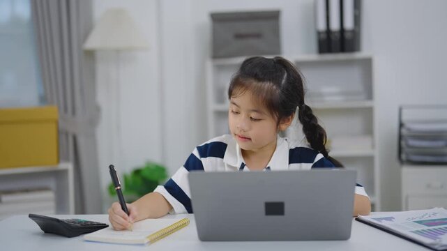 Young schoolgirl focused on studying homework using laptop and writing with pen at home desk in cozy room