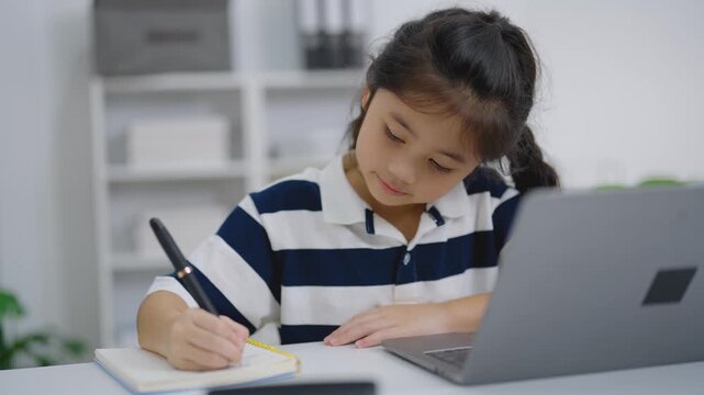 Young girl sitting at desk writing in notebook with pen while using laptop in bright modern study room at home