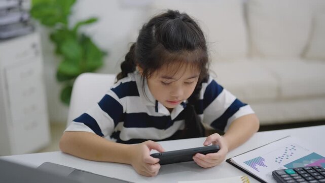 Young girl focused on tablet playing game or learning indoors at home sitting at white table with calculator