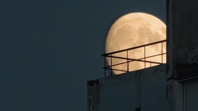 Full moon glowing behind metal railing on rooftop at night. Moon appears large behind weathered concrete structure with metal railing. Night sky dominates background. Urban nightscape