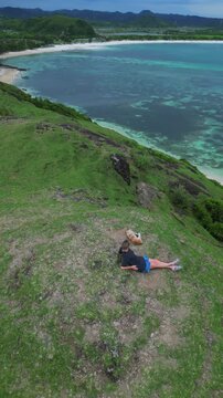 Woman Reclines On Cliff Beside Ocean, Solo Traveler Enjoying Panoramic Aerial View Of Turquoise Bay And Sandy Cove, Green Hills And Winding Shore In Background, Loyal Dog Curled Nearby, Lombok