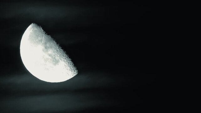 Moon appears in half phase with visible craters and texture, partially veiled by thin clouds. Cratered lunar surface of half moon glowing against dark night sky with wispy cloud cover