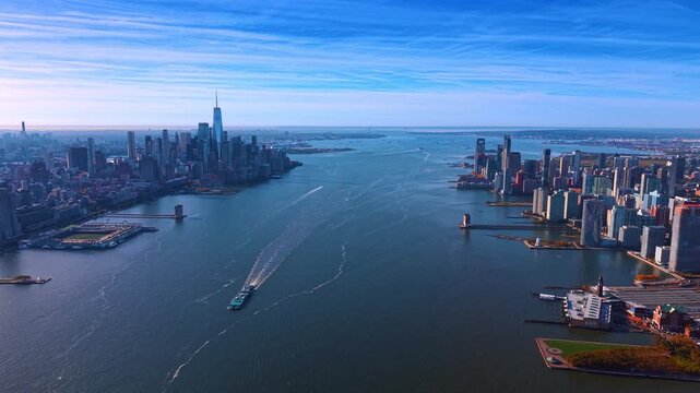 The Hudson River separates Manhattan and the New Jersey waterfront. Boats leave white trails between the two skylines with blue water widening across the foreground.