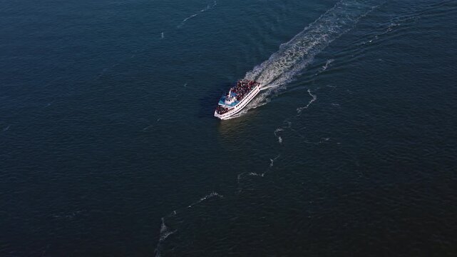 A passenger boat leaves a widening wake on the Hudson River. The white hull travels diagonally across dark blue water with people visible on the open upper deck.
