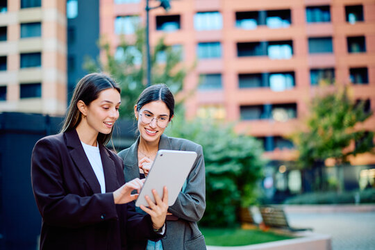 Two businesswomen collaborating over a tablet, sharing ideas and discussing work in an urban plaza surrounded by modern offices