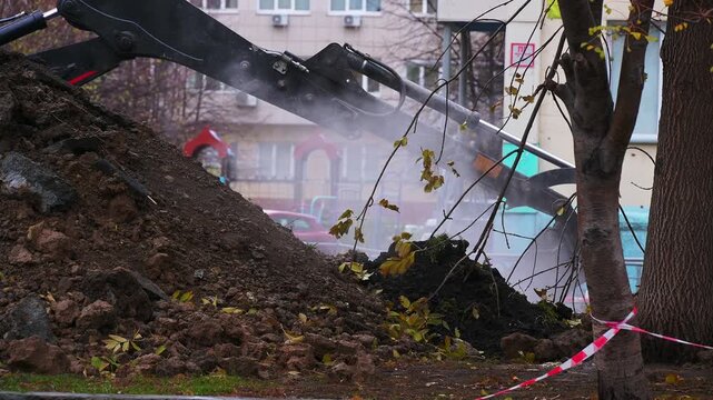 Urban excavation site with excavator arm, dirt pile, tree trunks, caution tape, playground in background, ongoing earthworks.
