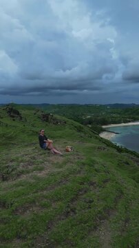 Caucasian woman sitting cliff ocean turquoise, holding dog on lap in panoramic aerial view of coral reef and sandy bay, dramatic cloud band on horizon, grassy headland, peaceful solitude, Lombok