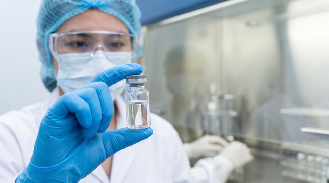 Focused scientist holds clear vaccine vial inside pharmaceutical laboratory. Laboratory worker wears blue sterile gloves while inspecting liquid medication beside sterile fume hood chamber.